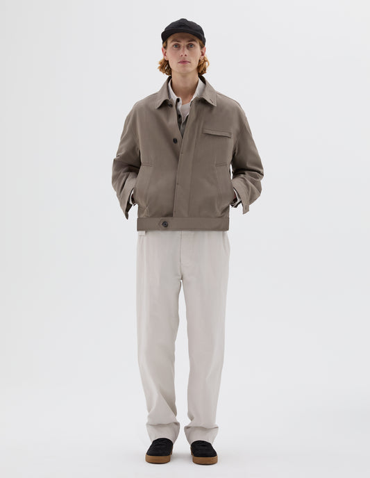 Full body image, front view of model wearing beige jacket with slant and chest pockets and horn button closure. Styled with light grey shirt, white t-shirt, off white trousers, black nubuck trainers and black cap.