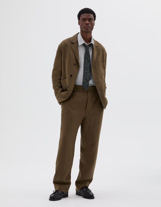 Full body image, front view of model wearing green/brown blazer with horn button closure and chest and patch pockets. Styled with light grey shirt, spotty green and beige tie, matching green/brown trousers and black leather boat shoes.