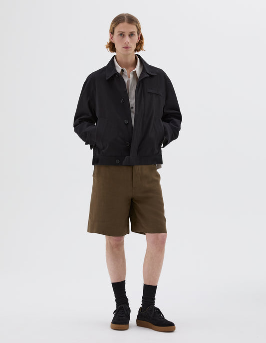 Full body image, front view of model wearing black jacket with slant and chest pockets and horn button closure. Styled with light grey shirt, green shorts, black socks and black nubuck trainers.