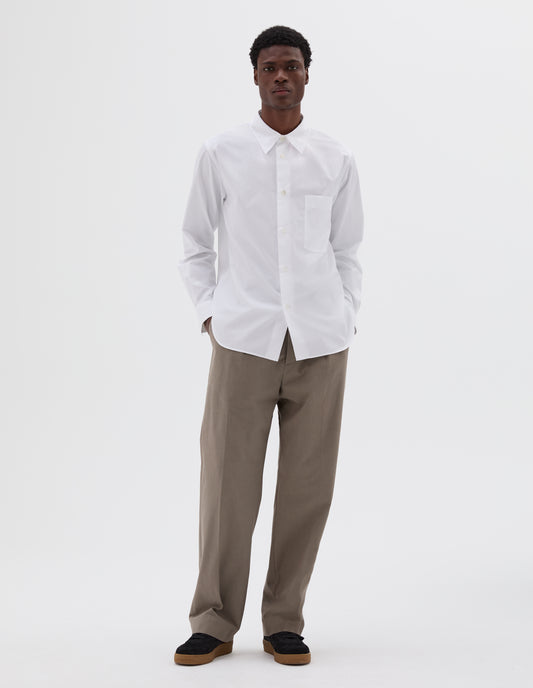 Full body image, front view of model wearing white cotton shirt with chest pocket, curved hem and mother of pearl buttons. Styled with beige trousers and black suede trainers.