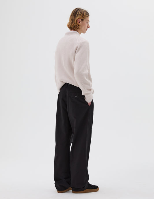 Full body image, back view of model wearing single pleated black trousers with button-through jet flap back pocket. Styled with white cotton shirt, off white ribbed jumper, black leather belt and black nubuck trainers.