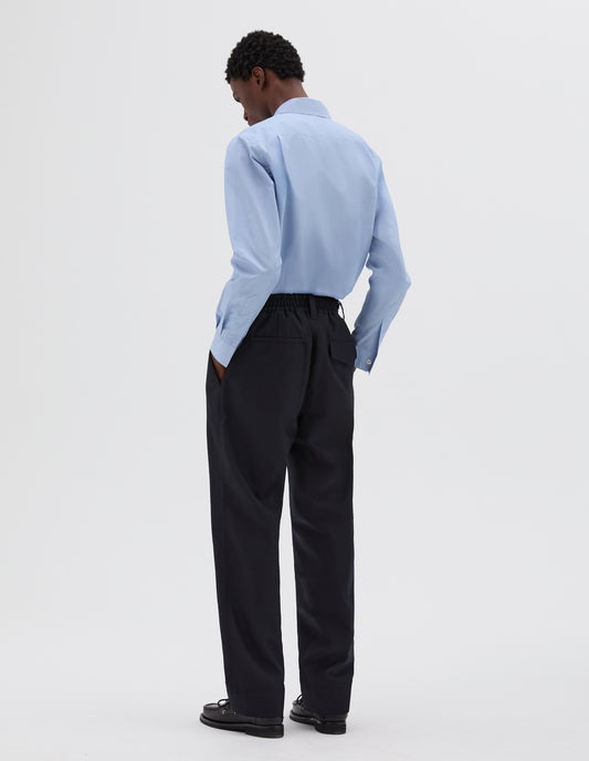 Full body image, back view of model wearing dark navy trousers, with half-elasticated waistband, belt loops and concealed button fastening flap back pockets. Styled with blue long sleeve shirt and black leather deck shoes.