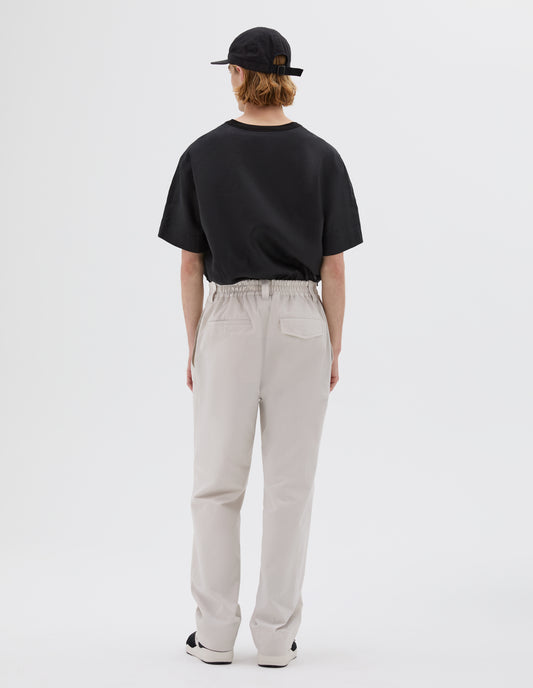 Full body image, back view of model wearing off white trousers, with half-elasticated waistband, belt loops and concealed button fastening flap back pockets. Styled with black woven t-shirt, black cap and black and brown trainers.