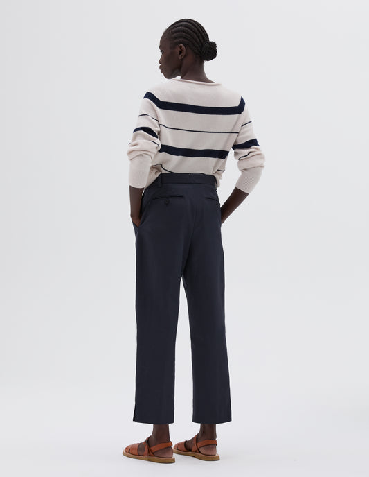 Full body image, back view of model wearing dark blue slim cut ankle crop trousers with midi waist, button-through welt back pocket and belt loops, styled with beige and blue stripy knitted jumper and light brown leather sandals.