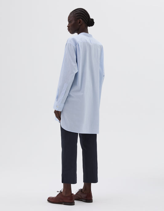 Full body image, back view of model wearing blue longline collarless shirt, with curved hem. Styled with cropped navy blue trousers and brown leather brogues.