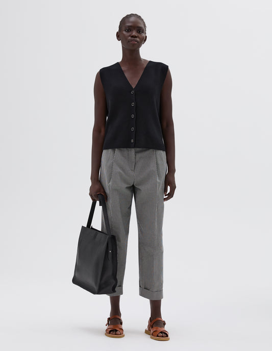 Full body image, front view of model wearing black boxy knitted waistcoat with horn buttons, styled with black and white gingham trousers with pleats, black leather tote and light brown leather sandals.