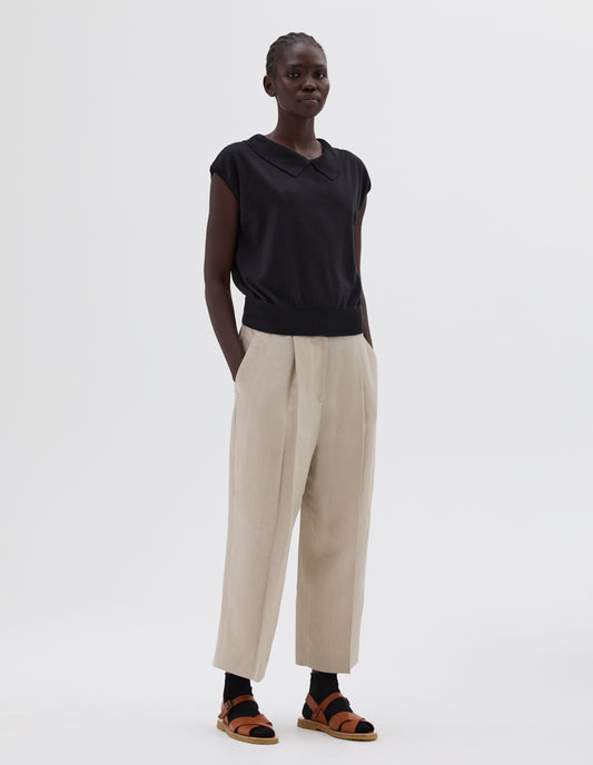 Full body image, front view of model wearing black relaxed collared knitted vest, styled with beige pleated trousers, black socks and light brown leather sandals.