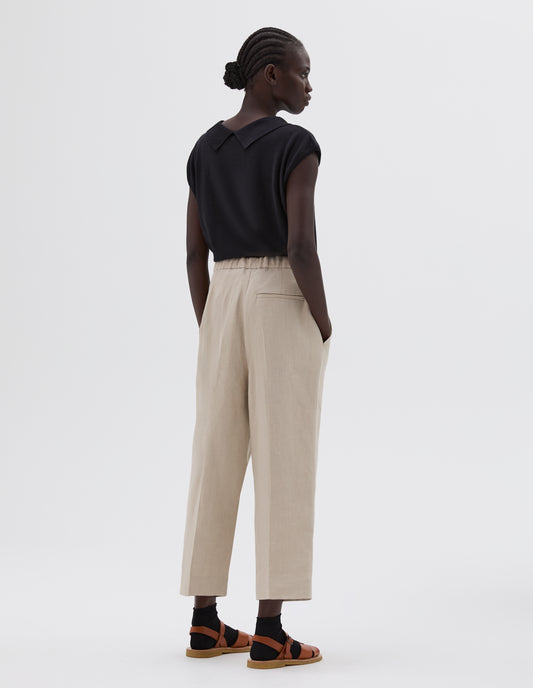 Full body image, back view of model wearing beige relaxed cut cropped length pleated trousers with side pockets, elasticated waist and welt back pocket, styled with black collared knitted vest and black socks with light brown leather sandals.