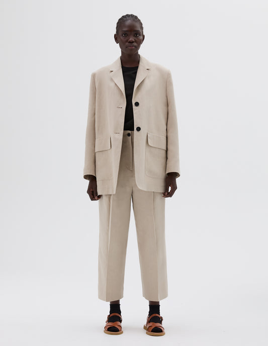 Full body image, front view of model wearing beige linen blazer with two front flap pockets and two horn button closure. Styled over a black round neck t-shirt, matching beige linen trousers, black ankle socks and light brown leather sandals. 