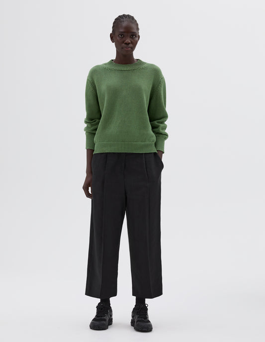 Full body image, front view of model wearing green relaxed chunky crew neck jumper, styled with black pleated trousers with side pockets, black socks and black trainers.