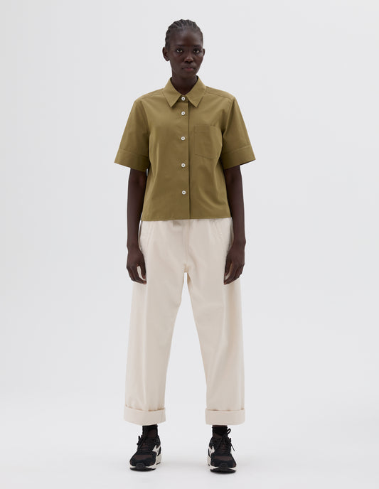 Full body image, front view of model wearing green short sleeve shirt with mother of pearl buttons and chest pocket. Styled with cropped white trousers, black socks and brown and black trainers.