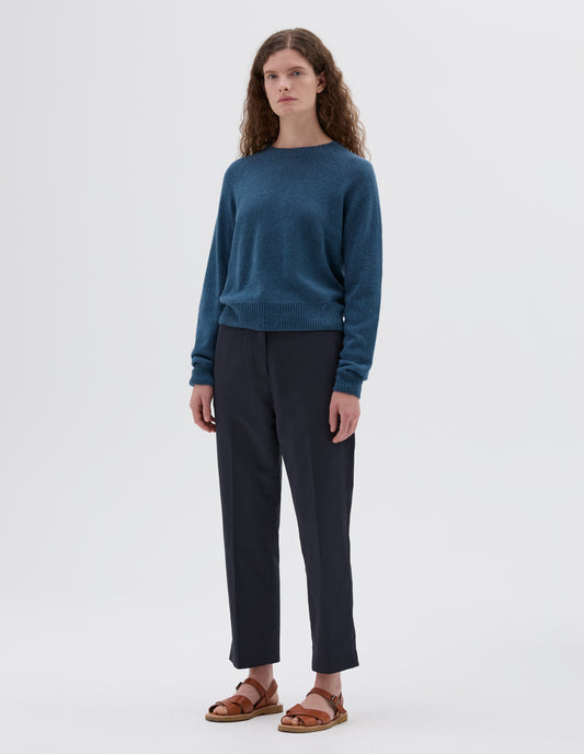 Full body image, front view of model wearing blue relaxed crew neck jumper with raglan sleeves, styled with dark blue cropped trousers and light brown leather sandals.