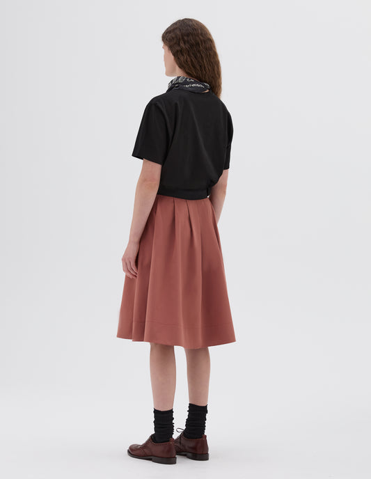 Full body image, back view of model wearing dark pink pleated midi length skirt with black waistband, styled with black t-shirt, black and white bandana, black socks with dark brown brogues.