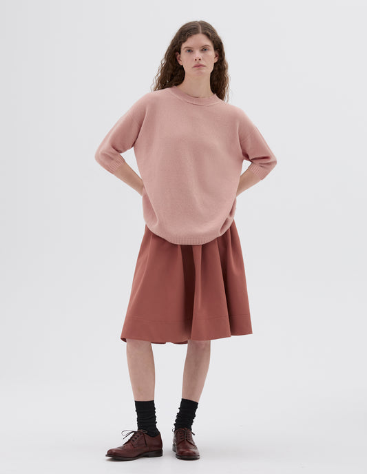 Full body image, front view of model wearing pink relaxed long length crew neck jumper with cropped sleeves, styled with dark pink skirt, black socks and dark brown brogues.