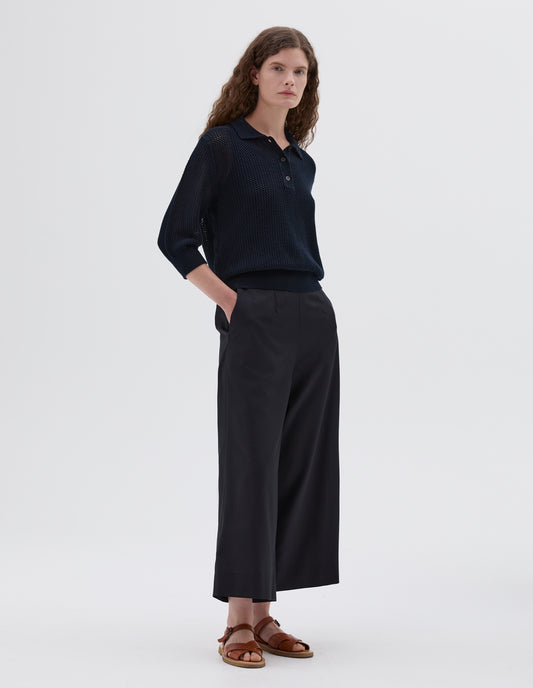Full body image, front view of model wearing dark blue relaxed pointelle polo shirt with mother of pearl buttons and cropped sleeves, styled with dark blue culottes with side pockets and light brown leather sandals.