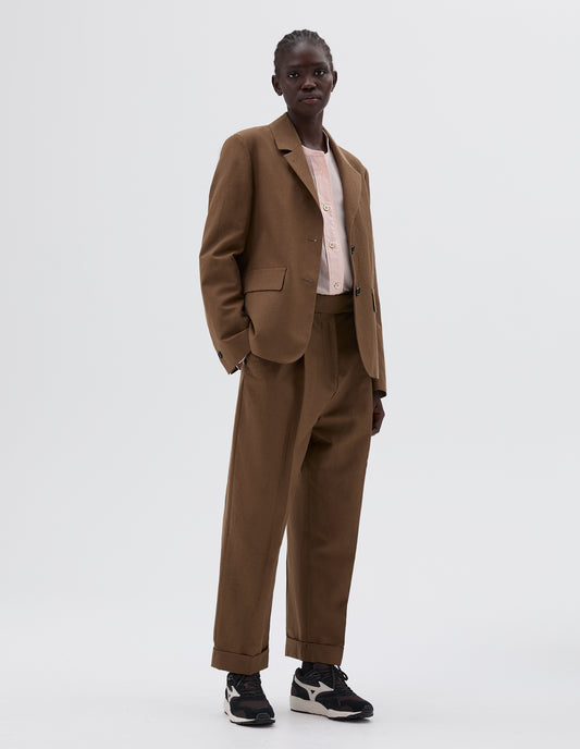 Full body image, front view of model wearing brown blazer with two front flap pockets and horn button closure, over a pink collarless blouse, Styled with matching brown trousers and brown and black trainers.