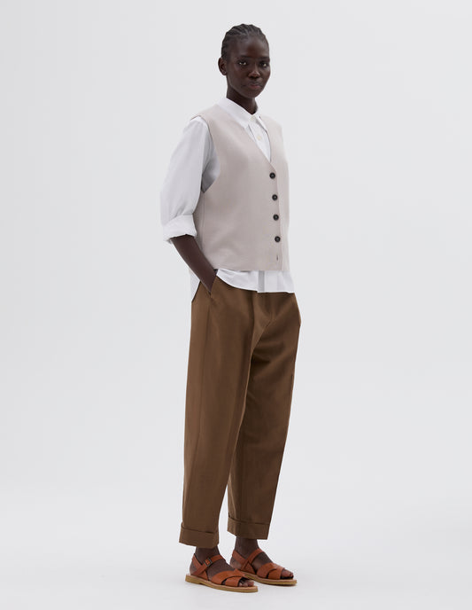 Full body image, side view of model wearing light grey boxy knitted waistcoat with black horn buttons, styled with white shirt and brown pleated trousers with side pockets and turned up hems, and light brown leather sandals.