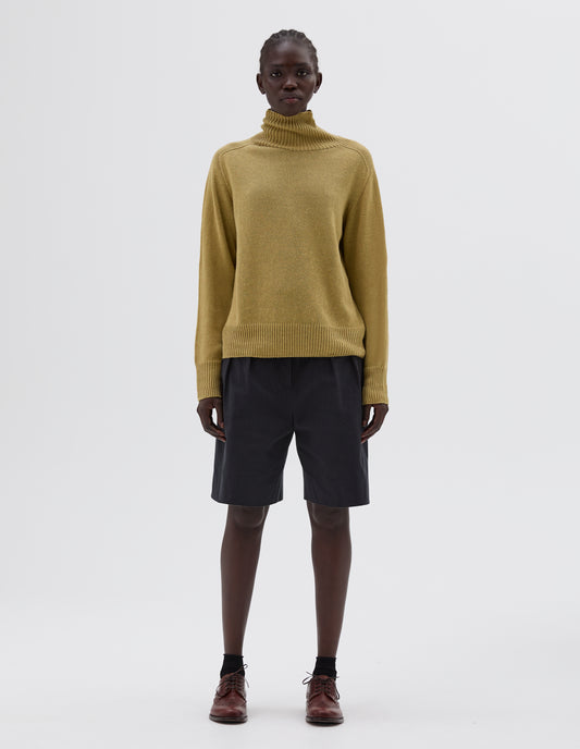 Full body image, front view of model wearing green relaxed roll neck jumper, styled with black pleated shorts with black socks and dark brown brogues.