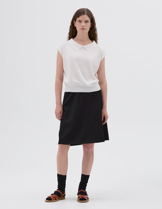 Full body image, front view of model wearing white relaxed collared knitted vest, styled with black skirt, black socks and light brown leather sandals.