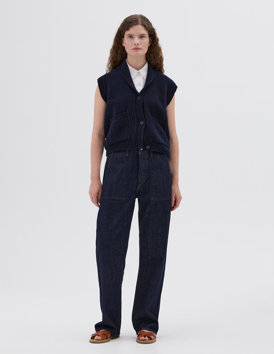 Full body image, front view of model wearing dark blue shawl collar knitted relaxed waistcoat with patch pockets and horn buttons, styled with white sleeveless collared shirt and dark blue jeans and light brown leather sandals.