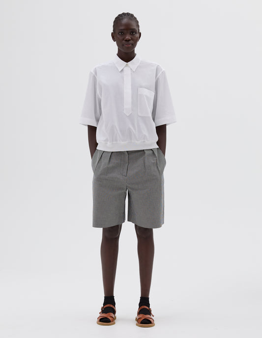 Full body image, front view of model wearing white shirt with chest pocket and contrasting elasticated hem. Styled with black and white gingham shorts, black socks and light brown leather sandals.