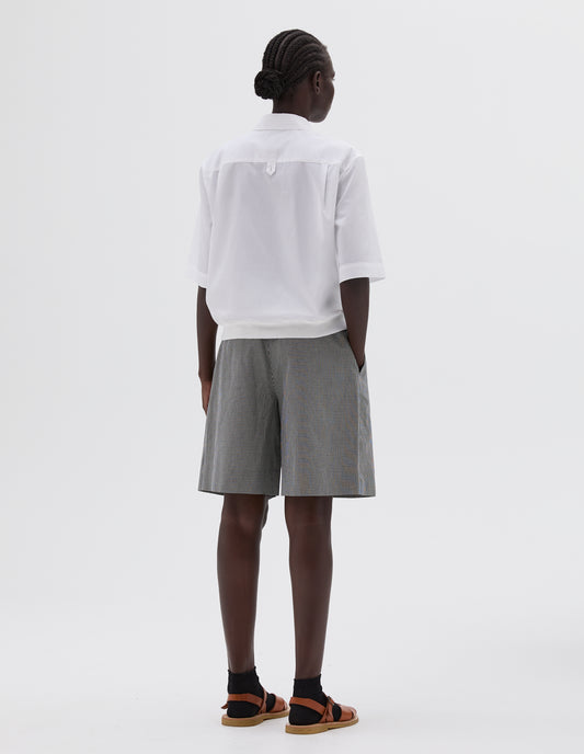 Full body image, back view of model wearing white shirt with contrasting elasticated hem and hanger loop on back yoke. Styled with black and white gingham shorts, black socks and light brown leather sandals.