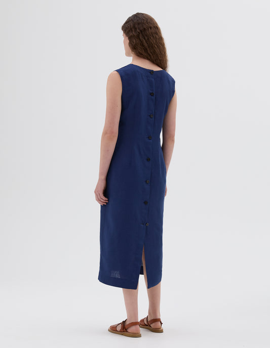 Full body image, back view of model wearing blue straight cut midi length sleeveless dress, horn buttons and deep back vent, styled with light brown leather sandals.