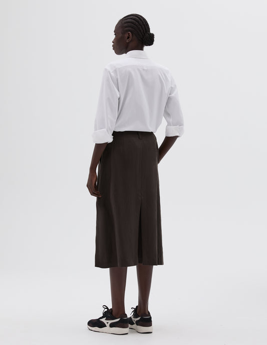 Full body image, back view of model wearing brown midi length skirt with back vent, belt loops and welt back pocket, styled with white long sleeve  collared shirt and black, brown and white lace up trainers.