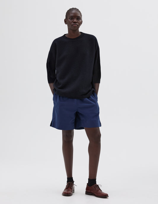 Full body image, front view of model wearing dark blue relaxed long length crew neck jumper with cropped sleeves, styled with blue shorts with side pockets and black socks with dark brown brogues. 