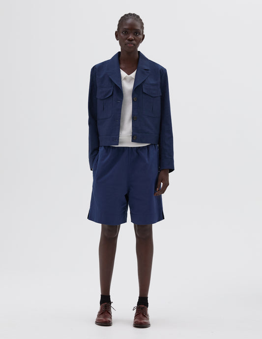 Full body image, front view of model wearing navy blue cropped utility jacket with two front flap pockets over a white knitted top. Styled with navy blue shorts, black socks and brown leather brogues. 