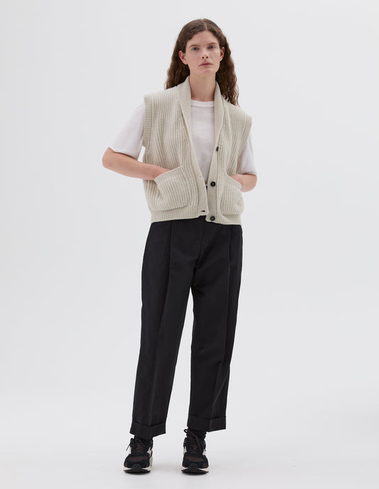 Full body image, front view of model wearing beige shawl collar knitted relaxed waistcoat with patch pockets and black horn buttons, styled with white t-shirt and black pleated trousers with turned up hems and black, brown and white trainers.
