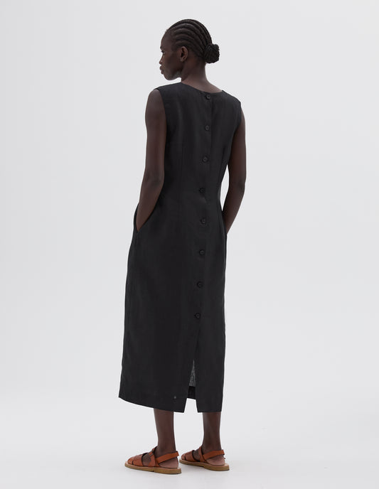 Full body image, back view of model wearing black straight cut midi length sleeveless dress with side pockets, horn buttons and deep back vent, styled with light brown leather sandals.