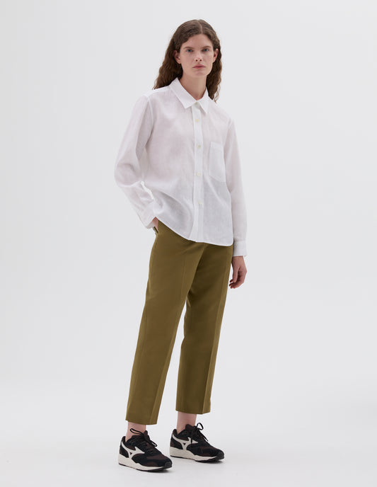 Full body image, front view of model wearing white linen shirt, with mother of pearl buttons and chest pocket. Styled with cropped green trousers and brown and black trainers.