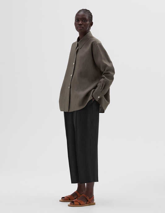 Full body image, front view of model wearing brown long sleeve shirt, with mother of pearl button closure and cuffs. Styled with cropped black trousers and light brown leather sandals.