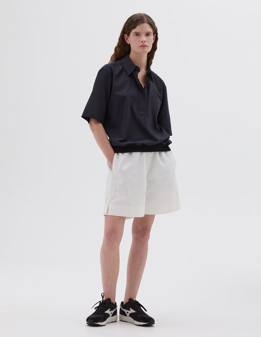 Full body image, front view of model wearing black shirt with chest pocket and contrasting elasticated hem. Styled with white shorts and brown and black trainers. 