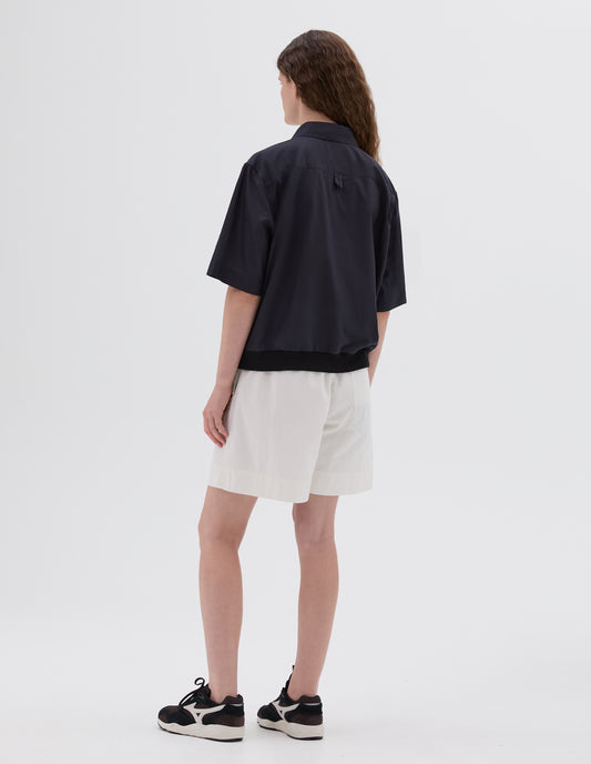 Full body image, back view of model wearing black shirt with contrasting elasticated hem and hanger loop on back yoke. Styled with white shorts and brown and black trainers. 