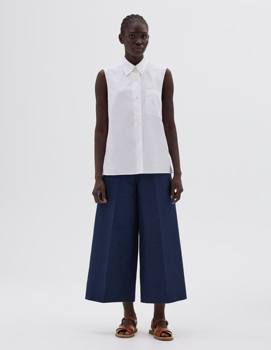 Full body image, front view of model wearing white sleeveless shirt, with mother of pearl button and chest pocket. Styled with blue culottes and light brown leather sandals. 