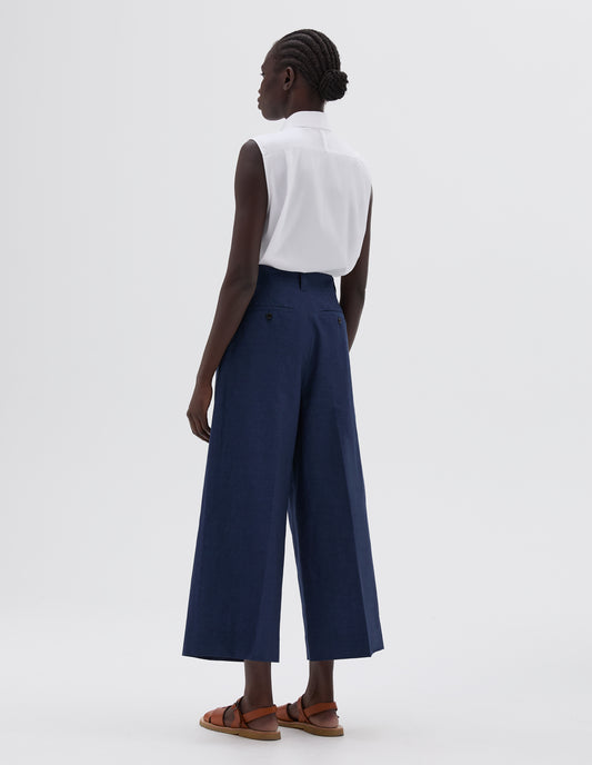 Full body image, back view of model wearing blue culottes with button-through welt back pockets and belt loop, styled with white sleeveless collared shirt and light brown leather sandals.