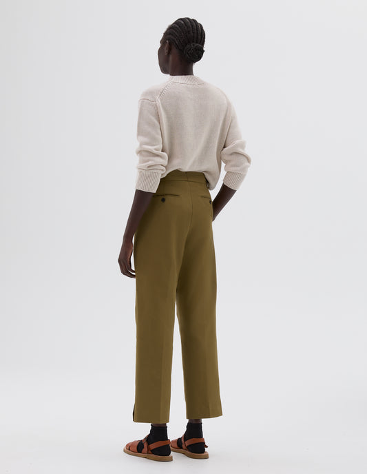 Full body image, back view of model wearing green slim cut ankle crop trousers with midi waist, button-through welt back pocket and belt loops, styled with beige knitted jumper, black socks and light brown leather sandals.