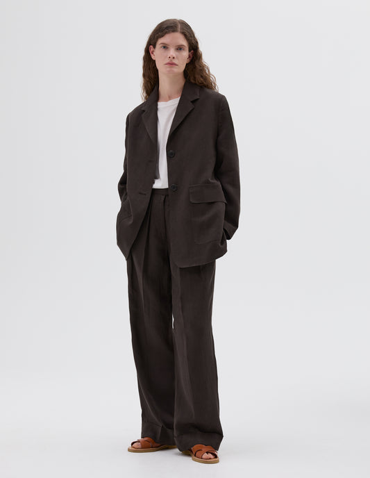 Full body image, front view of model wearing brown linen blazer with two front flap pockets and horn button closure. Styled over a white round neck t-shirt, with matching brown linen trousers and light brown leather sandals. 