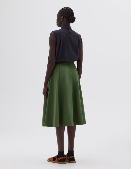 Full body image, back view of model wearing green midi length flared skirt, styled with black sleeveless collared shirt, black socks and light brown leather sandals.