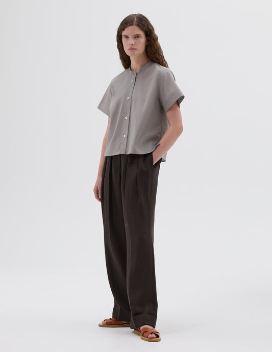 Full body image, front view of model wearing grey collarless short sleeve shirt, with mother of pearl buttons. Styled with brown linen trousers and light brown leather sandals. 