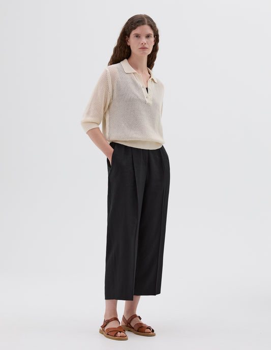 Full body image, front view of model wearing beige relaxed pointelle polo shirt with mother of pearl buttons and cropped sleeves, styled with black vest and black pleated trousers with side pockets and light brown leather sandals.