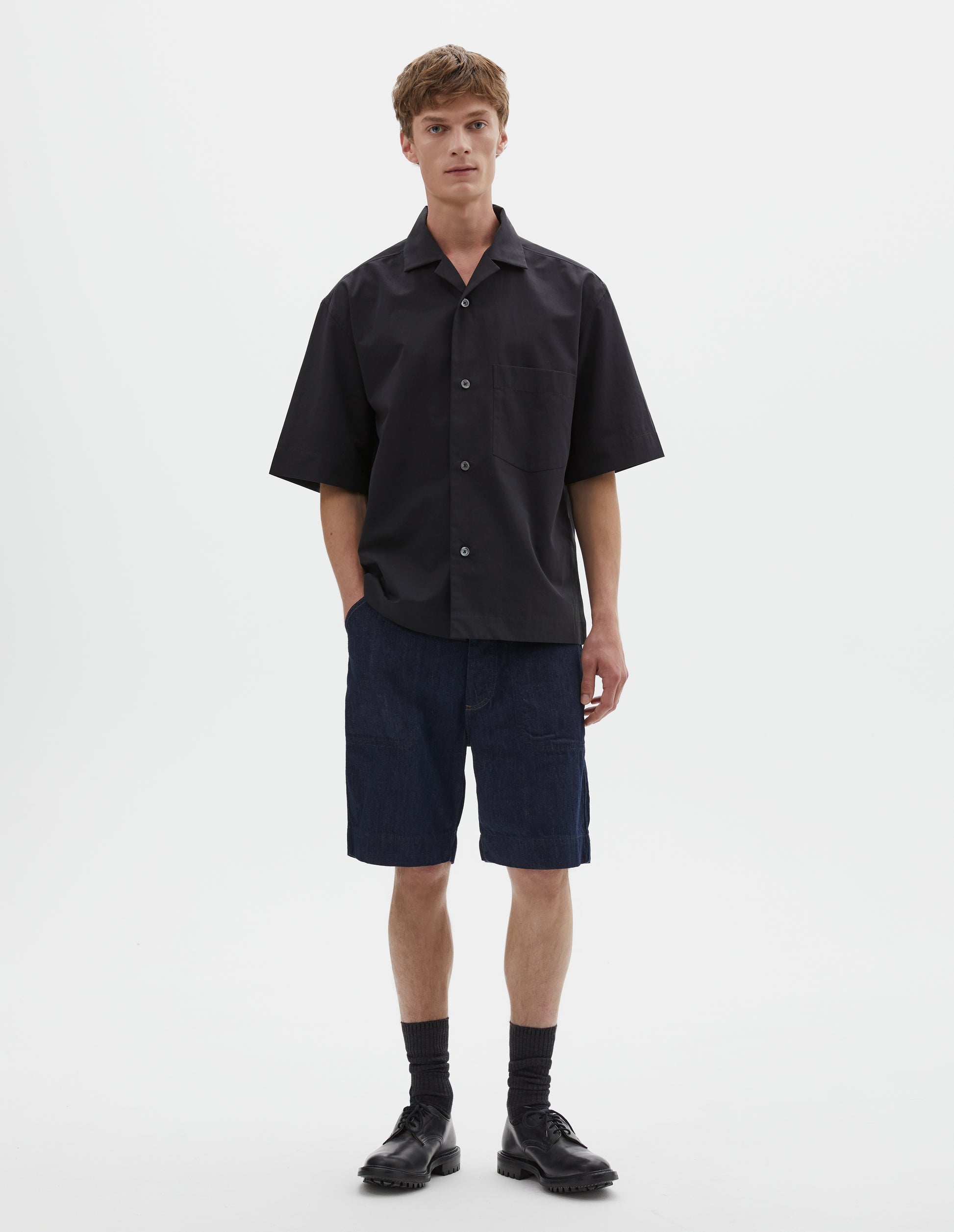 Full body image, front view of model wearing relaxed boxy short-sleeved black shirt with revere collar, mother of pearl buttons, chest patch pocket and side split hem. Styled with dark blue denim shorts, black socks and black leather shoes. 