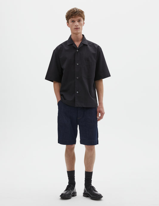 Full body image, front view of model wearing relaxed boxy short-sleeved black shirt with revere collar, mother of pearl buttons, chest patch pocket and side split hem. Styled with dark blue denim shorts, black socks and black leather shoes. 