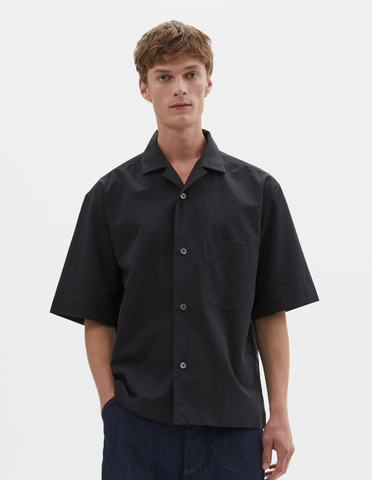Cropped image, front view of model wearing relaxed boxy short-sleeved black shirt with revere collar, mother of pearl buttons, chest patch pocket and side split hem. Styled with dark blue denim shorts. 