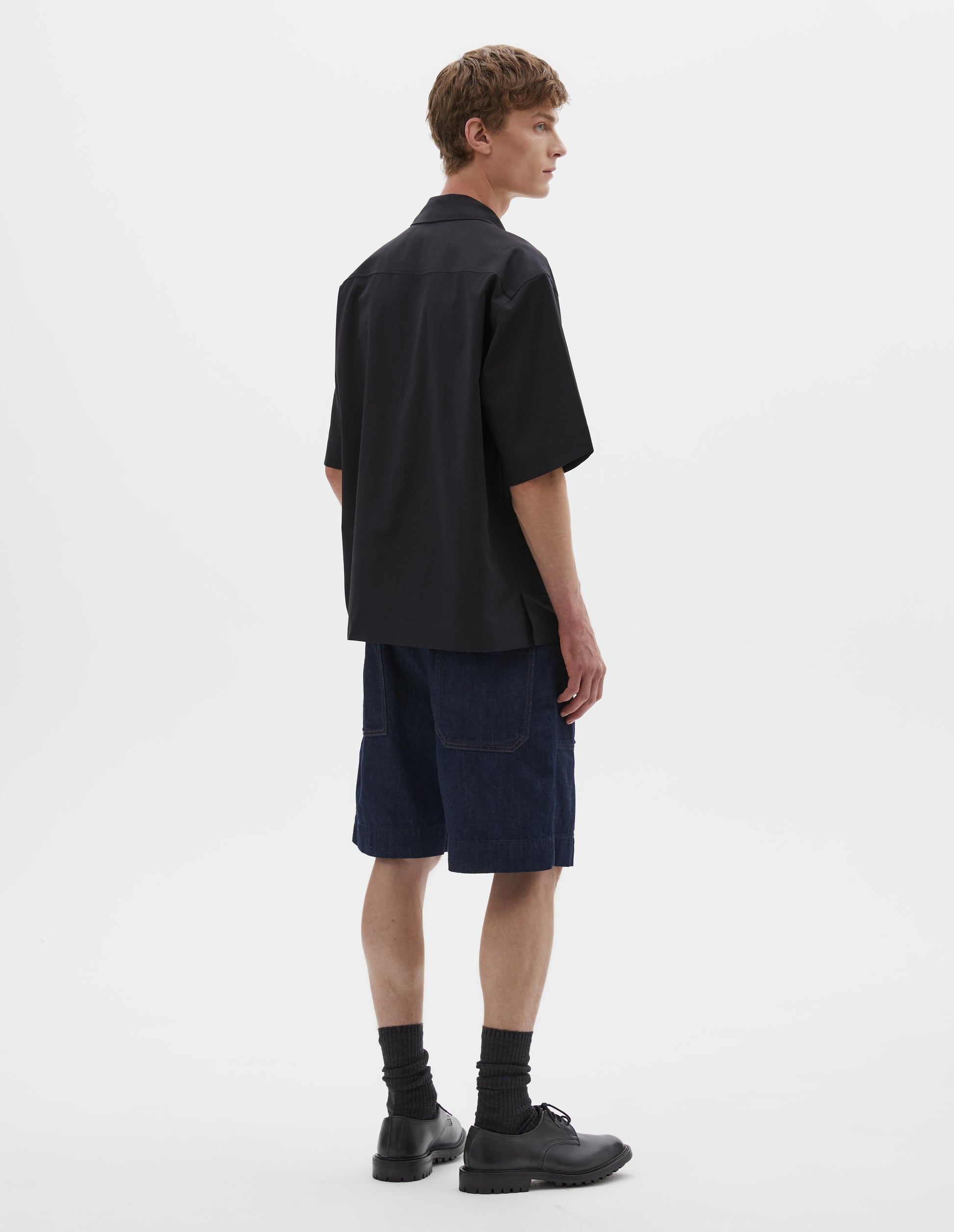 Full body image, back view of model wearing relaxed boxy short-sleeved black shirt with side split hem. Styled with dark blue denim shorts, black shoes and black leather shoes. 
