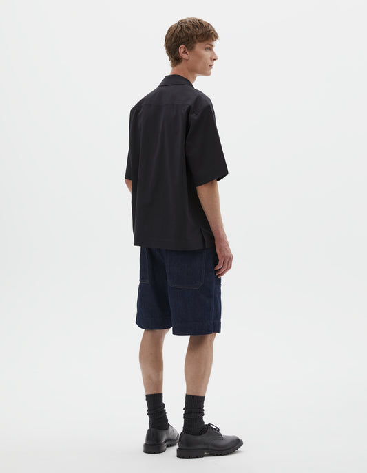 Full body image, back view of model wearing relaxed boxy short-sleeved black shirt with side split hem. Styled with dark blue denim shorts, black shoes and black leather shoes. 
