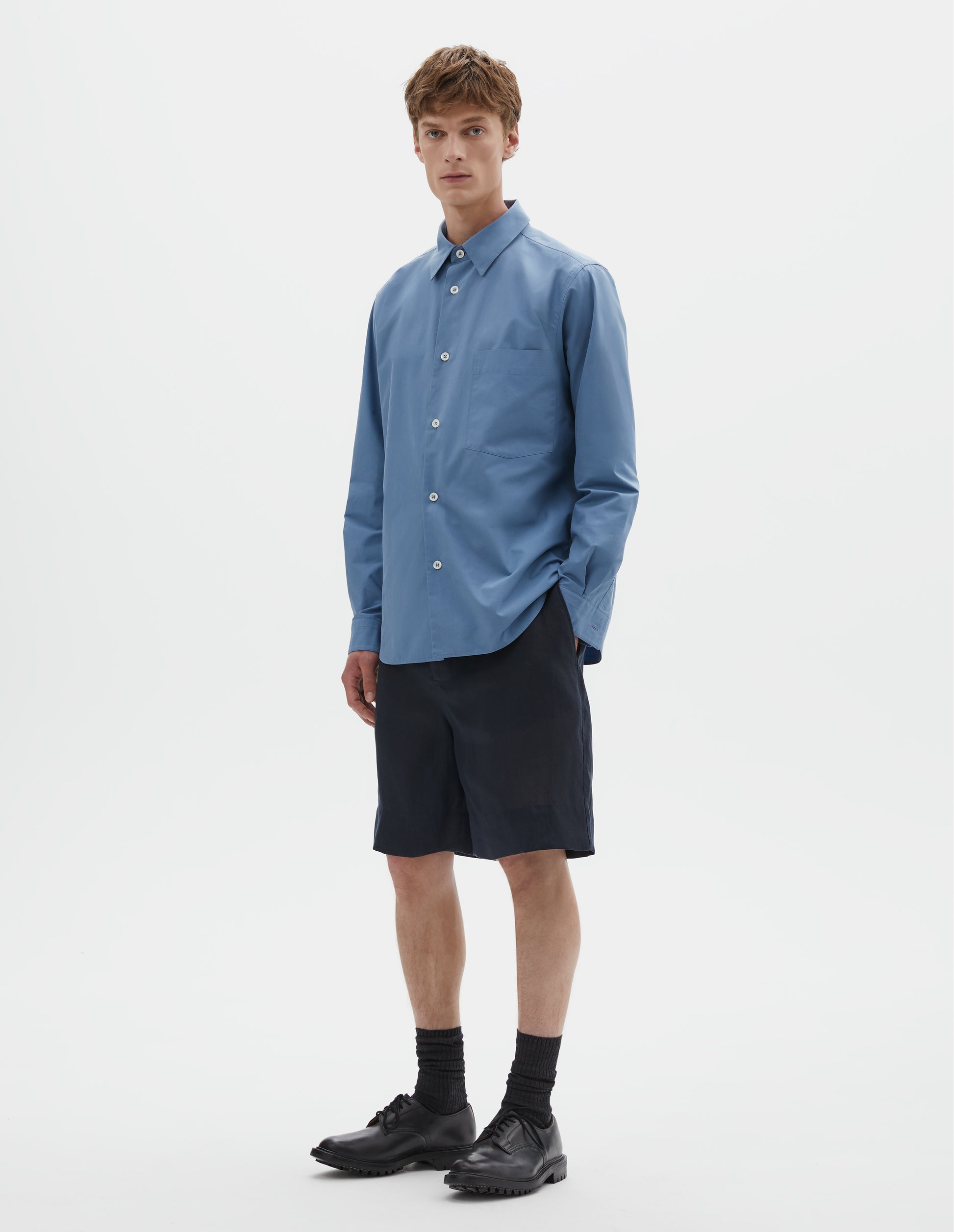 Full body image, front view of model wearing blue cotton long sleeve shirt, with mother of pearl buttons, curved hem and chest patch pocket. Styled with dark navy blue shorts, black socks and black leather brogues.