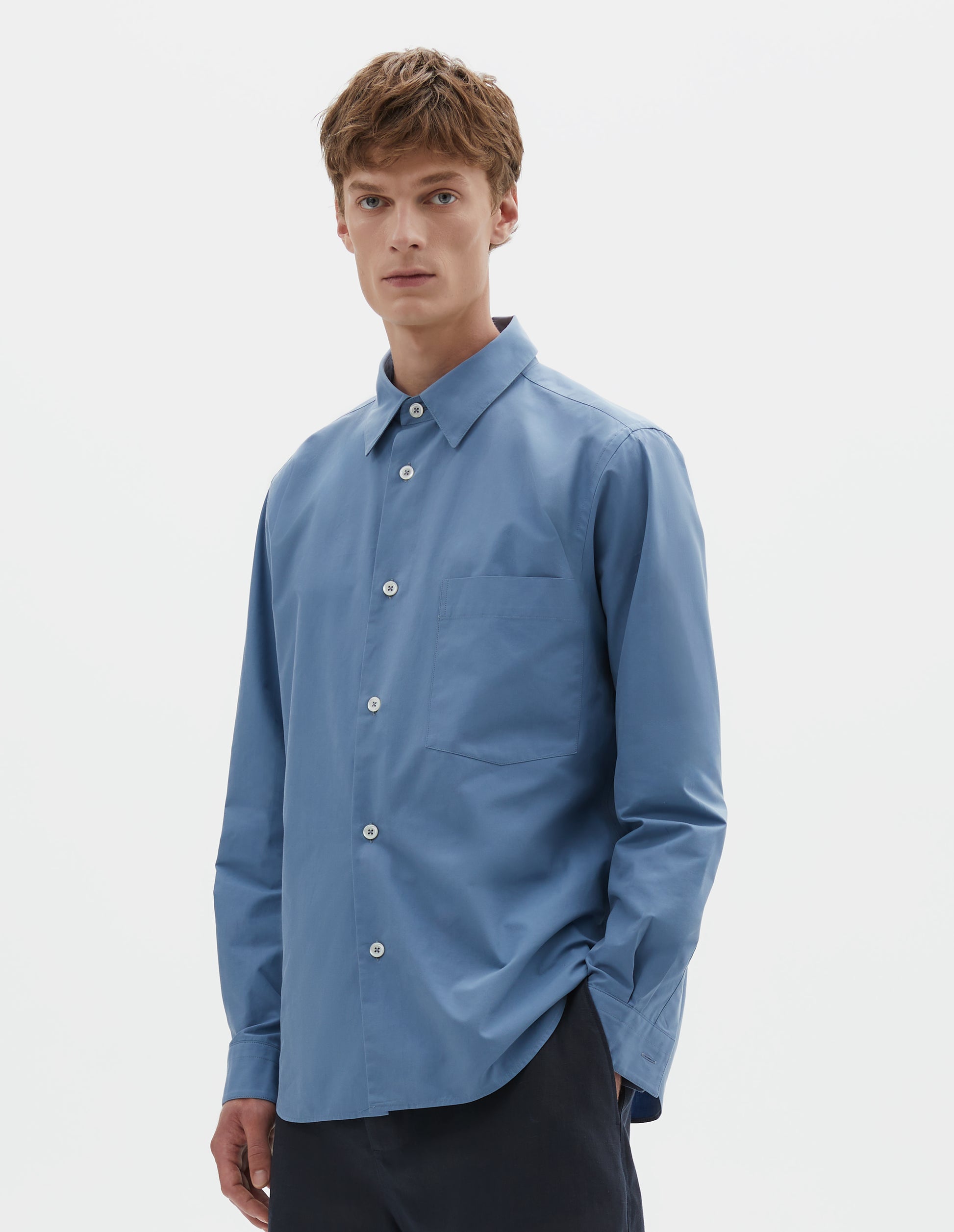 Cropped body image, front view of model wearing blue cotton long sleeve shirt, with mother of pearl buttons, curved hem and chest patch pocket. Styled with dark navy blue shorts.